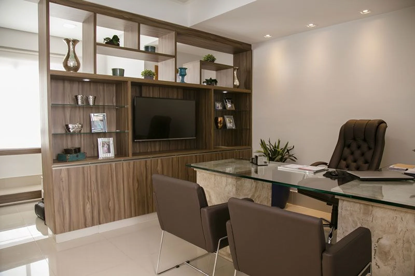 Sala de consulta: mesa de vidro sobre base de mármore, duas cadeiras de couro marrom, poltrona capitonê e estante de madeira com objetos