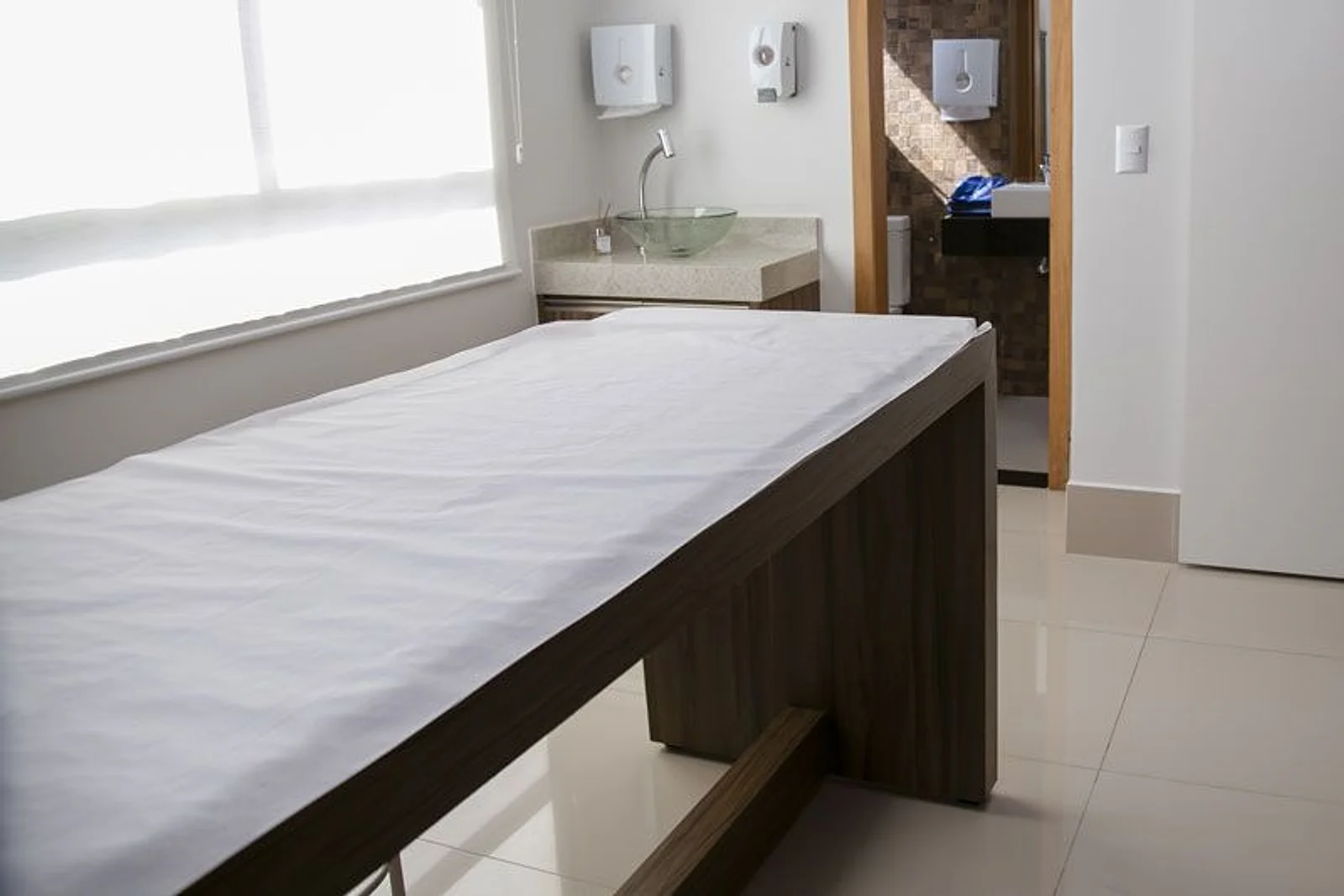 Sala de exame: maca de madeira forrada com lençol branco, pia de vidro junto à janela e porta para o banheiro