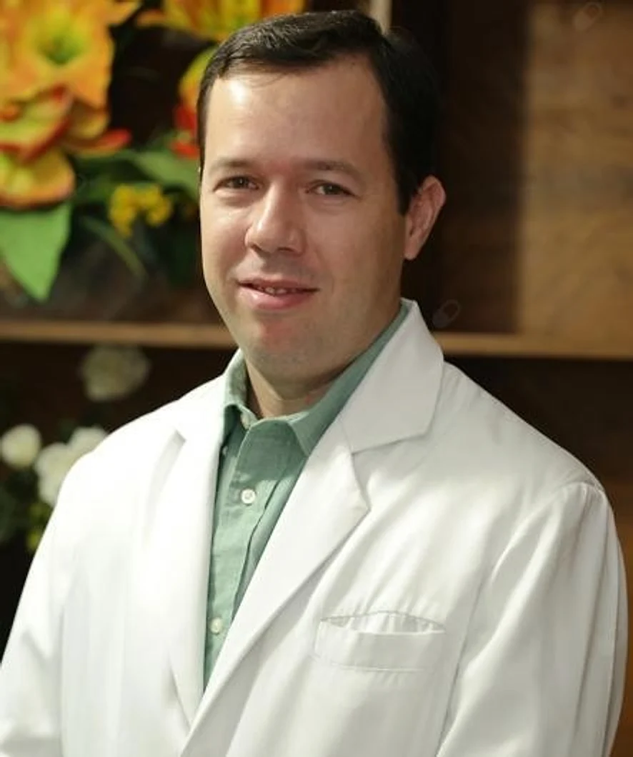 Dr. Fabricio Leão de jaleco branco e camisa verde, sorrindo, com flores amarelas e madeira do consultório ao fundo