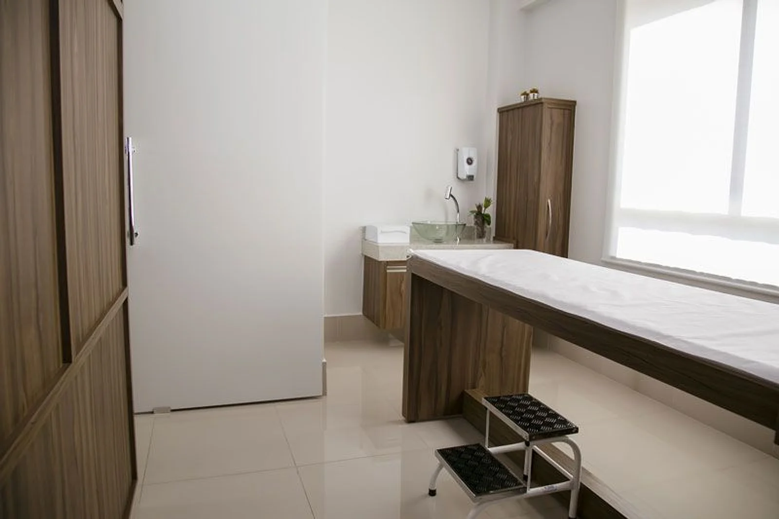 Sala de exame do consultório: maca de madeira com lençol branco, escadinha, pia e armário de madeira, luz de janela
